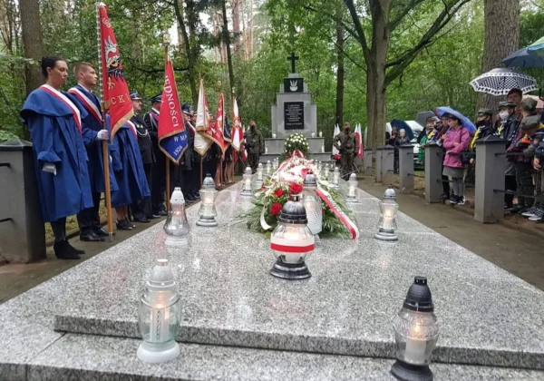 Powiatowa uroczystość upamiętniająca wybuch II wojny światowej, z uczestnikami w strojach ceremonialnych trzymającymi flagi po lewej, żołnierzami po prawej oraz kwiatami i zapalonymi lampionami na granitowej płycie pomnika.
