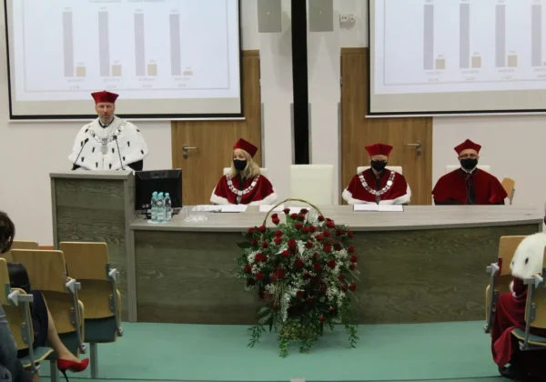 Sala konferencyjna podczas inauguracji Roku Akademickiego 2020/2021 w płockich uczelniach: osoba w białej todze przemawia z mównicy, za nią siedzą trzy osoby w czerwonych togach, przed nimi duża kwiatowa dekoracja, a w tle projekcje na ekranach.