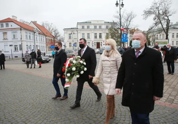 Grupa osób w maseczkach idzie ulicą podczas obchodów Narodowego Święta Niepodległości; jeden z mężczyzn niesie biało‑czerwony wieniec z kwiatów.