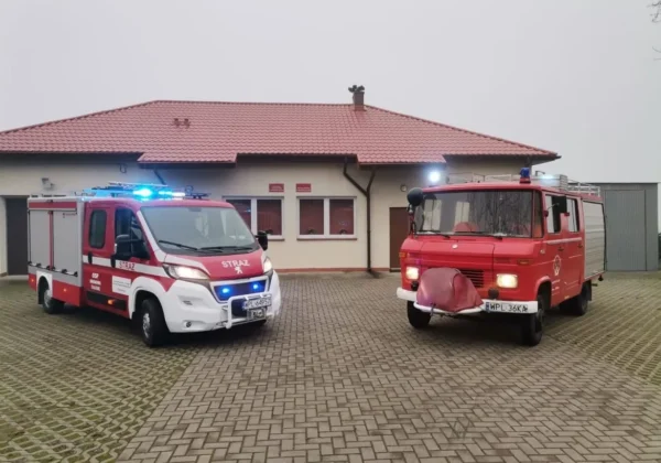 Dwa wozy straży pożarnej stoją przed remizą: nowoczesny biało‑czerwony samochód z niebieskimi światłami po lewej i starszy czerwony wóz gaśniczy po prawej, na tle budynku z czerwonym dachem.