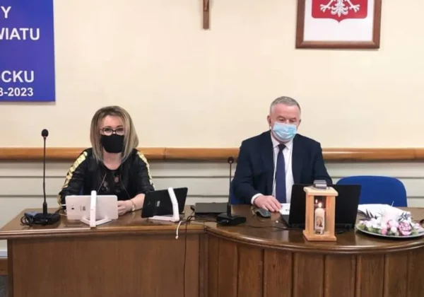Dwóch urzędników siedzi przy biurku podczas XXI sesji Rady Powiatu w Płocku; oboje w maseczkach, przed nimi mikrofony i laptopy, a w tle widoczny krzyż i godło Polski.
