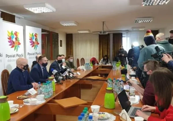 Posiedzenie sztabu kryzysowego w Powiecie Płockim z udziałem służb odpowiedzialnych za bezpieczeństwo powodziowe, przy stole konferencyjnym z mikrofonami, kamerami i dziennikarzami.