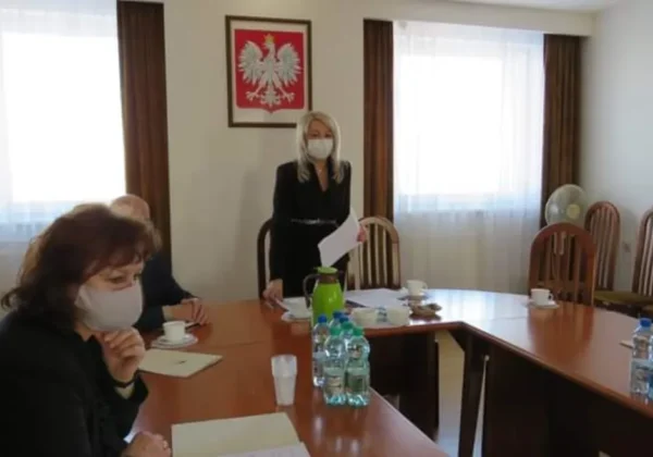 Uczestnicy Powiatowej Rady Rynku Pracy w maseczkach podczas posiedzenia w sali konferencyjnej, przy stole z dokumentami, napojami i butelkami wody, na tle godła państwowego.