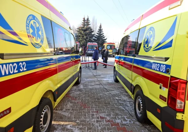 Dwa nowoczesne ambulanse w żółtej kolorystyce z czerwonymi pasami stoją na brukowanym placu w Wyszogrodzie, a w tle widać barierkę i kilka osób.