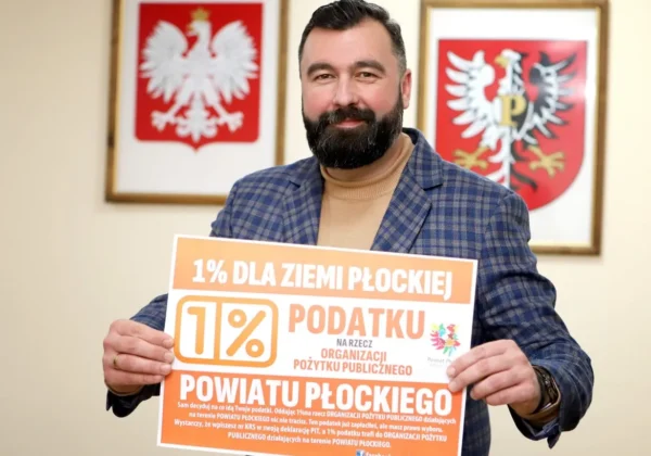Mężczyzna z brodą w kratkowanej marynarce trzyma plakat promujący 1% podatku dla Ziemi Płockiej, stojąc w biurze z herbami w tle.