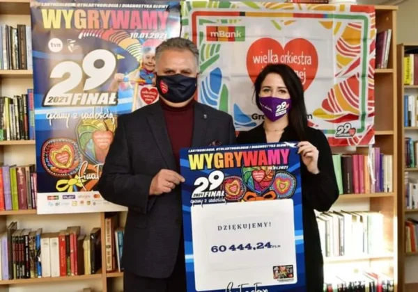 Dwoje osób w maseczkach stoi w bibliotecznej przestrzeni obok banerów WOŚP i trzyma duży plakat z kwotą 60 444,24 zł.