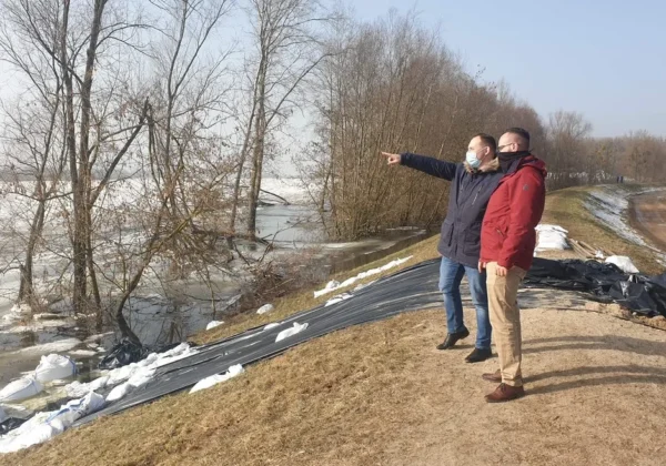 Dwóch mężczyzn w kurtkach i maseczkach stoi na brzegu rzeki, wskazując na prowadzone działania zabezpieczenia przeciwpowodziowego, obok czarne plandeki i worki z piaskiem.