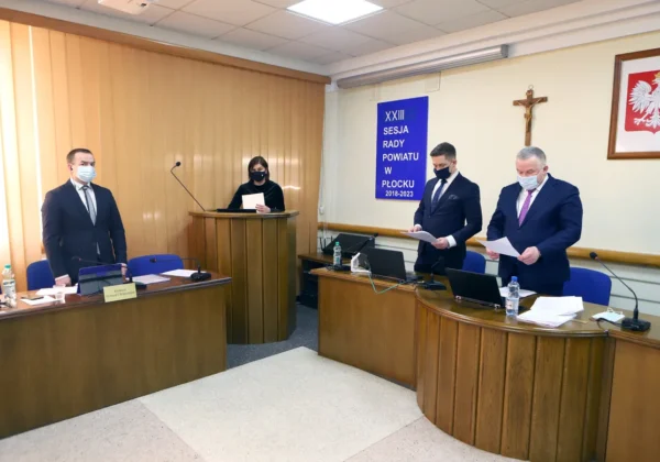 Czterech urzędników w maseczkach podczas XXIII Sesji Rady Powiatu w Płocku stoi przy drewnianych stanowiskach z laptopami i dokumentami; na ścianie wisi baner z napisem XXIII Sesja Rady Powiatu w Płocku 2018–2023, obok krzyż i godło Polski.