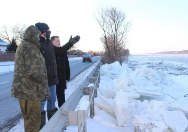 Trójka osób w zimowych ubraniach stoi przy barierce nad zamarzniętą rzeką, obserwując drogę i czujnie zwracając uwagę na niebezpieczeństwo lodu.