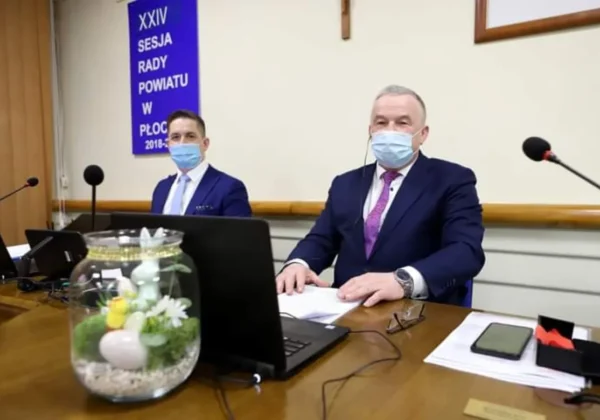 Dwóch mężczyzn w garniturach i maseczkach siedzi przy stole w sali obrad podczas XXIV sesji Rady Powiatu w Płocku, z laptopami i mikrofonami.
