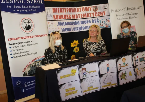 Trzy kobiety w maseczkach pracują przy stole z laptopami podczas finału Konkursu Matematycznego w Zespole Szkół im. Jana Śniadeckiego w Wyszogrodzie, na tle banerów i plakatów promujących konkurs.