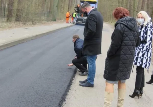 Grupa ludzi obserwuje postęp prac przy budowie nowej drogi w Powiecie Płockim; na pierwszym planie mężczyzna klęczy przy krawędzi świeżo położonej nawierzchni, w tle widać maszynę drogową.
