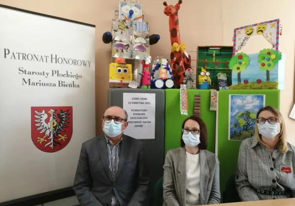 Trójka osób w maseczkach siedzi przy stole podczas Powiatowego Konkursu Ekologicznego z okazji Międzynarodowego Dnia Ziemi, na tle kolorowych dekoracji i plakatów promujących wydarzenie.