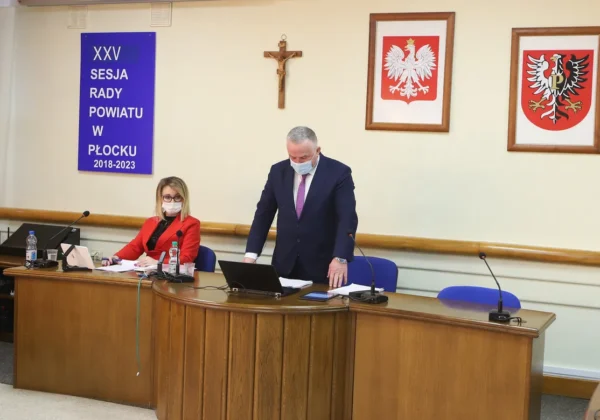 XXV sesja Rady Powiatu w Płocku: Zarząd Powiatu przy stole sesyjnym, kobieta w czerwonym żakiecie pracuje na laptopie, a mężczyzna w garniturze omawia dokumenty; w tle widoczne są krzyż i dwa herby.