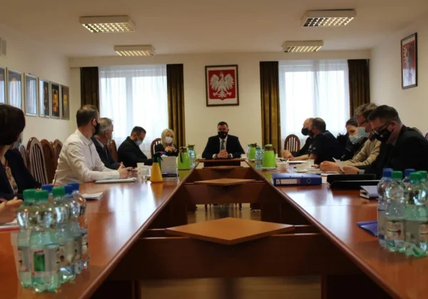 Grupa osób w maseczkach siedzi przy długim stole konferencyjnym podczas posiedzenia Powiatowego Zespołu Zarządzania Kryzysowego ws. ptasiej grypy, z centralnie siedzącym prowadzącym i godłem Polski na ścianie w tle.