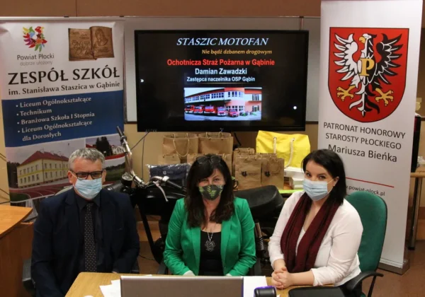 Trzy osoby w maseczkach siedzą przy stole przed ekranem z prezentacją i bannerami Powiatu Płockiego oraz Staszic MotoFan.