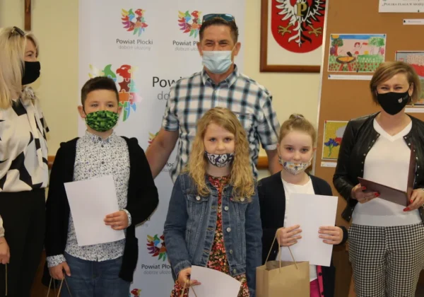 Grupa dorosłych i dzieci podczas rozdania nagród – wszyscy w maseczkach, trzymają dyplomy i upominki przed tłem z logami Powiatu Płockiego.