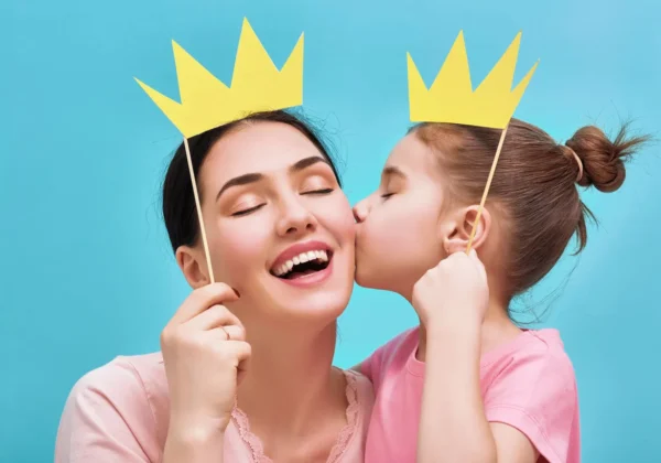 Uśmiechnięta mama trzyma żółtą papierową koronę na patyku, a jej córka całuje ją w policzek, oboje stoją na jasno-niebieskim tle.