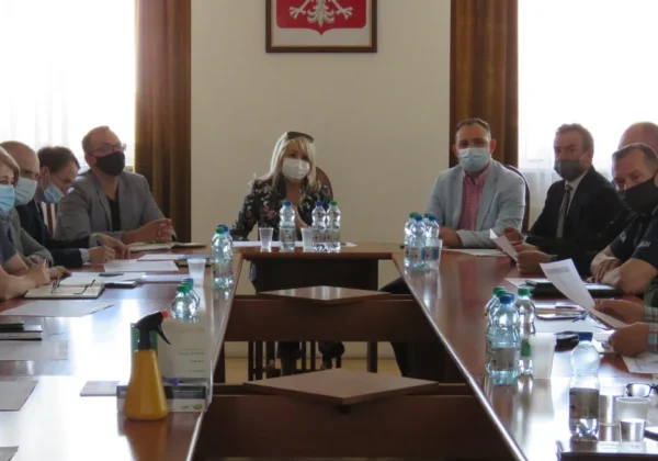 Grupa osób w maseczkach siedzi przy długim stole w sali konferencyjnej, omawiając dokumenty podczas spotkania dotyczącego ogniska ptasiej grypy w powiecie płockim.