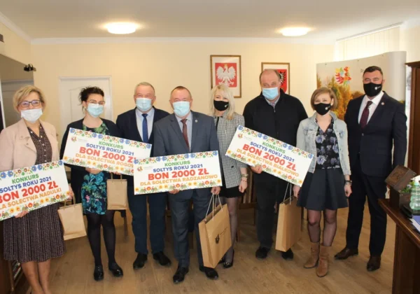 Grupa osób w maseczkach stoi w biurze urzędu i prezentuje duże, kolorowe certyfikaty z nagrodami „Sołtys Roku 2021” (bon 2000 zł dla sołectw).