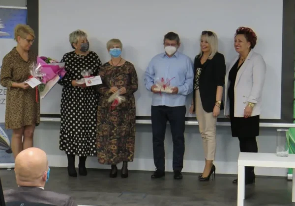 Grupa sześciu osób stoi na scenie podczas wydarzenia w bibliotece, niektórzy mają maseczki, trzymając kwiaty i upominki, a w tle widać kamerę na statywie i banery.