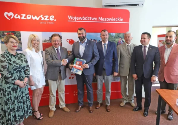 Grupa siedmiu osób w garniturach i eleganckich strojach stoi przed czerwonym banerem z napisem Mazowsze – serce Polski i Województwo Mazowieckie; w centrum dwie osoby ściskają dłonie, trzymając stos dokumentów.