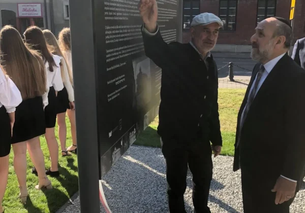 Dwóch mężczyzn stoi przy czarnej tablicy upamiętniającej żydowską społeczność Gąbina, z których jeden gestem wskazuje tekst, a za nim widać grupę dziewcząt w białych bluzkach i czerwono‑białą wstęgę.