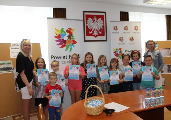 Grupa dzieci wraz z opiekunkami odbiera nagrody i dyplomy podczas konkursu „Lubię spędzać czas ze swoją rodziną” w Powiecie Płockim, stojąc przed bannerem z herbem powiatu.