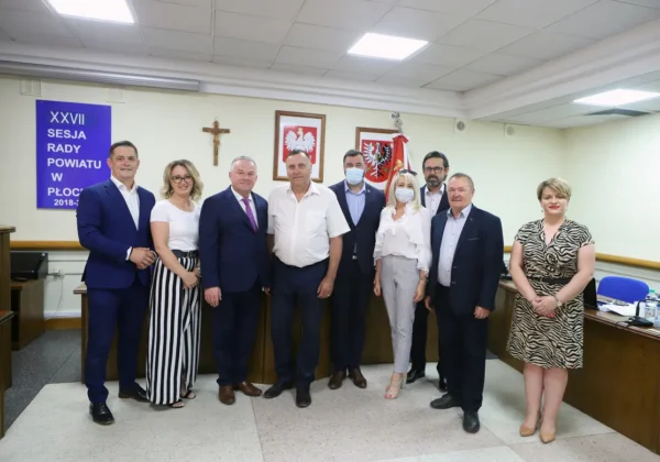 Grupa urzędników w formalnych ubraniach stoi w sali sesyjnej Rady Powiatu w Płocku podczas XXVII sesji; nowy radny złożył ślubowanie.