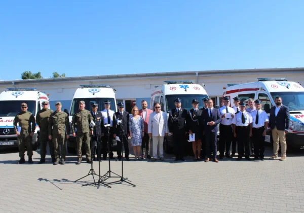 Grupa osób w mundurach strażackich, policyjnych i strojach cywilnych stoi przed szeregiem ambulansów podczas uroczystości dla strażaków-ochotników z Powiatu Płockiego.