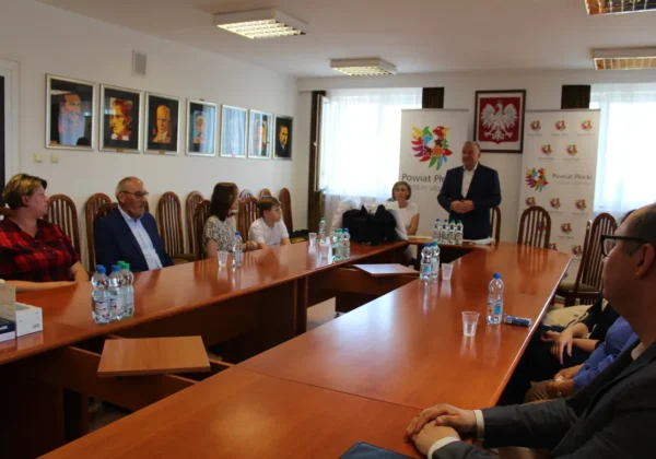 Grupa osób siedzi przy długim stole w sali konferencyjnej; jeden mężczyzna stoi i wygłasza przemówienie podczas wręczania nagród autorom wierszy o pszczołach.