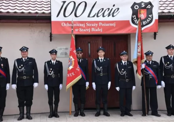 Grupa strażaków Ochotniczej Straży Pożarnej w Starej Białej stoi w mundurach z białymi rękawiczkami przed wejściem do budynku, trzymając flagi i banner upamiętniający 100-lecie jednostki.