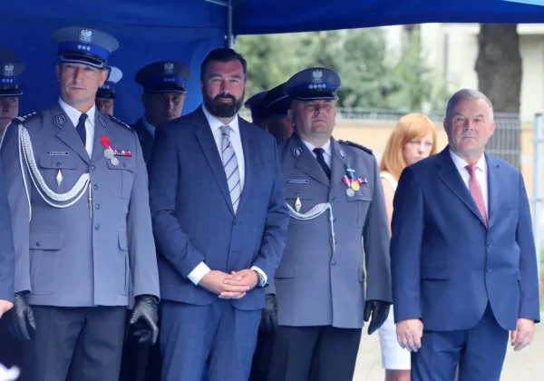 Grupa czterech osób, dwóch policjantów w szarych mundurach z odznaczeniami i dwóch mężczyzn w garniturach, stoi pod niebieskim namiotem podczas Święta Policji, gdzie wręczane są medale i upominki dla funkcjonariuszy od władz Powiatu Płockiego.