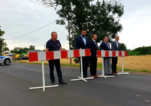 Pięciu mężczyzn stoi za czerwono-białymi barierami na świeżo wyremontowanej drodze w gminie Radzanowo, obok radiowozu policji i pól w tle.