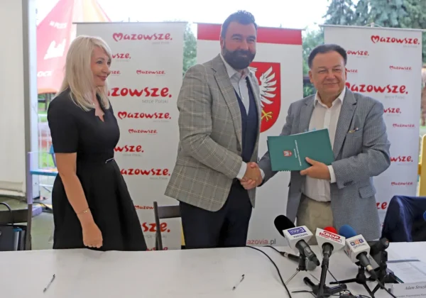 Trzy osoby stojące przy stole z mikrofonami podczas konferencji prasowej, dwaj mężczyźni ściskają dłonie, obok nich kobieta, w tle banery Mazowsze i zapowiedź nowoczesnego obiektu sportowego przy szkole w Wyszogrodzie.