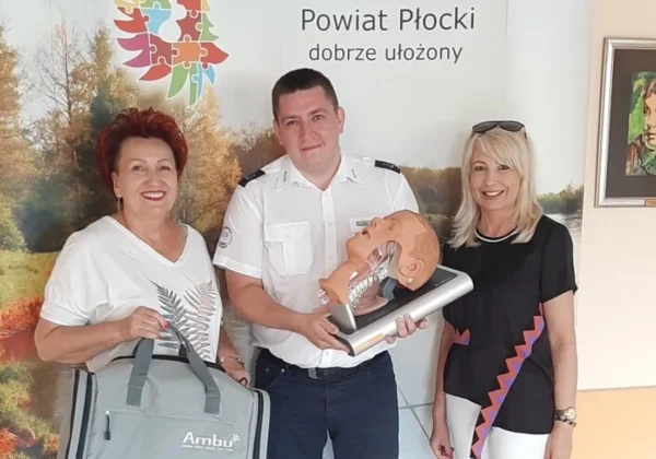 Trzy osoby stojące na tle baneru Powiatu Płockiego, a mężczyzna w mundurze trzyma szkoleniowy manekin do nauki udrażniania dróg oddechowych i resuscytacji.