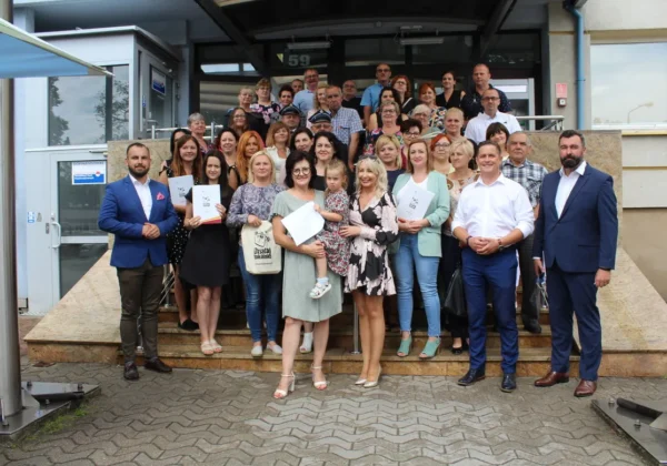 Grupa mieszkańców i samorządowców stoi na schodach przed budynkiem urzędu, trzymając dokumenty i symbolicznie świętując granty dla swoich gmin.