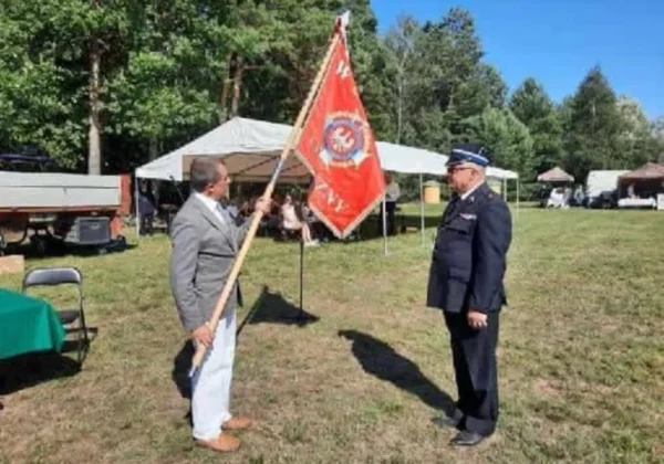 Mężczyzna w garniturze trzyma czerwony sztandar z emblematem przed strażakiem w mundurze podczas uroczystości Stulecia OSP Żerniki, na tle namiotów i zielonego placu.