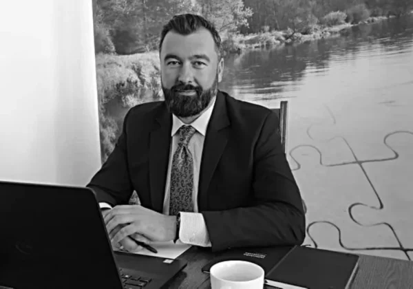 Czarno-białe zdjęcie mężczyzny w garniturze siedzącego przy biurku z laptopem, kubkiem i notatnikiem, w tle widoczny mural z motywem puzzli i krajobrazu.