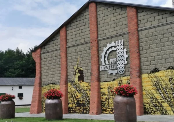 Ściana budynku z muralem przedstawiającym zboże i logo PGR Koszelew, a przed nim stoją trzy duże naczynia z czerwonymi kwiatami.
