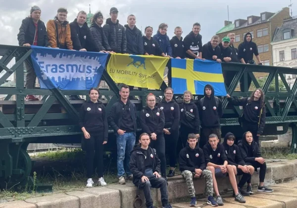 Grupa młodych ludzi stoi na moście, prezentując banery Erasmus+, EU Mobility Sweden i flagę Szwecji, podczas kończących się praktyk.