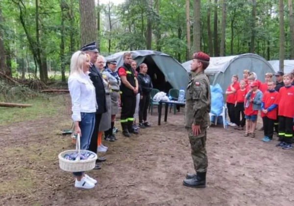 Zakończenie obozu szkoleniowo-wypoczynkowego dla Młodzieżowych Drużyn Pożarniczych: grupa młodych strażaków w czerwonych koszulkach stoi w lesie przed namiotami, słuchając przemówienia prowadzącego w mundurze.