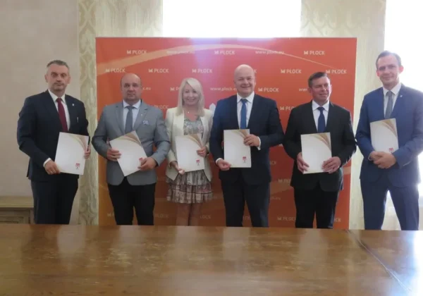 Sześciu urzędników i samorządowców stoi w sali konferencyjnej przed banerem z logiem Płocka, trzymając podpisane dokumenty, symbolizujące współpracę Powiatu Płockiego w Obszarze Funkcjonalnym Miasta Płocka.