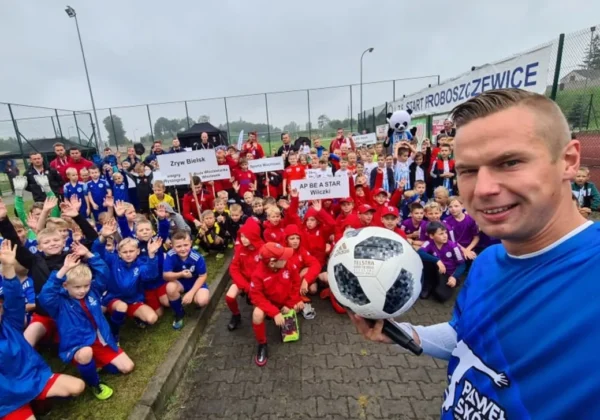 Grupa młodych piłkarzy w kolorowych strojach zgromadzona na boisku w Proboszczewicach, trener w niebieskiej koszulce trzyma piłkę i robi selfie na tle banerów i ogrodzenia.