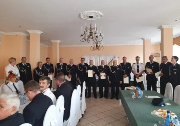 Grupa druhów Ochotniczej Straży Pożarnej w Słupnie stoi w rzędzie w sali podczas zjazdu, trzymając dokumenty, na tle bankietowej sali z żyrandolem i stołem.