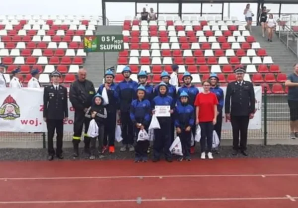 Grupa młodzieżowych drużyn pożarniczych w niebieskich kombinezonach wraz z opiekunami stoi na bieżni stadionu podczas X Wojewódzkich Zawodów Sportowo-Pożarniczych w Białobrzegach.