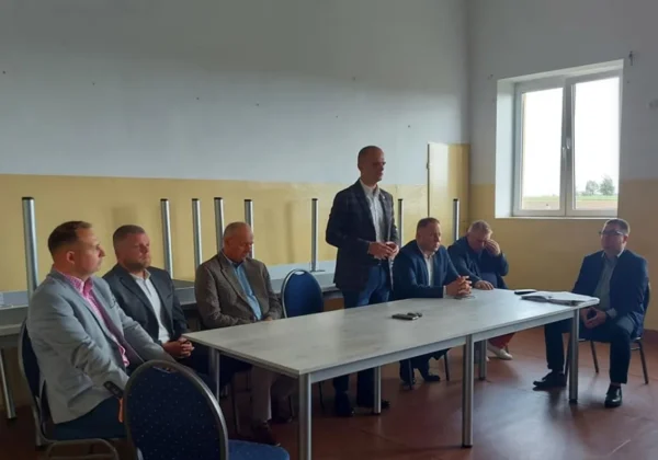 Mężczyzna stoi i przemawia przy stole podczas spotkania z mieszkańcami gminy Bulkowo, podczas gdy pozostali mężczyźni siedzą i słuchają w sali.