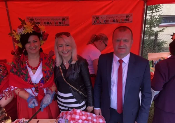 Trzy osoby stoją przy stole z ciastem podczas parafialnego święta plonów w Dobrzykowie, wśród czerwonego namiotu: kobieta w kwiecistym, tradycyjnym nakryciu głowy i czerwonej sukni, druga w czarnym skórzanym żakiecie, a mężczyzna w garniturze z czerwonym krawatem.