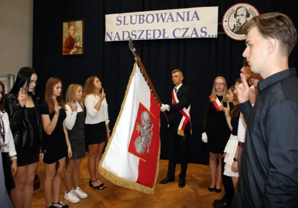 Grupa uczniów ubranych formalnie stoi w rzędzie i składa przysięgę podczas uroczystości ślubowania klas pierwszych, obok flagi państwowej na tle banneru.
