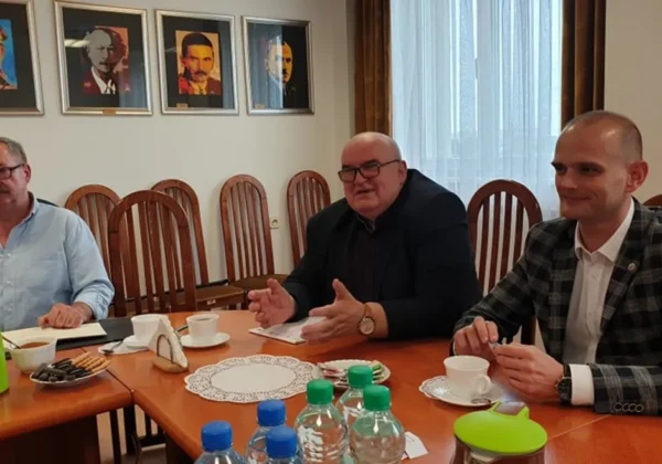 Trzech mężczyzn siedzi przy długim stole w sali konferencyjnej podczas spotkania; na stole znajdują się filiżanki, talerzyki z przekąskami i butelki wody, a w tle wiszą kolorowe portrety.